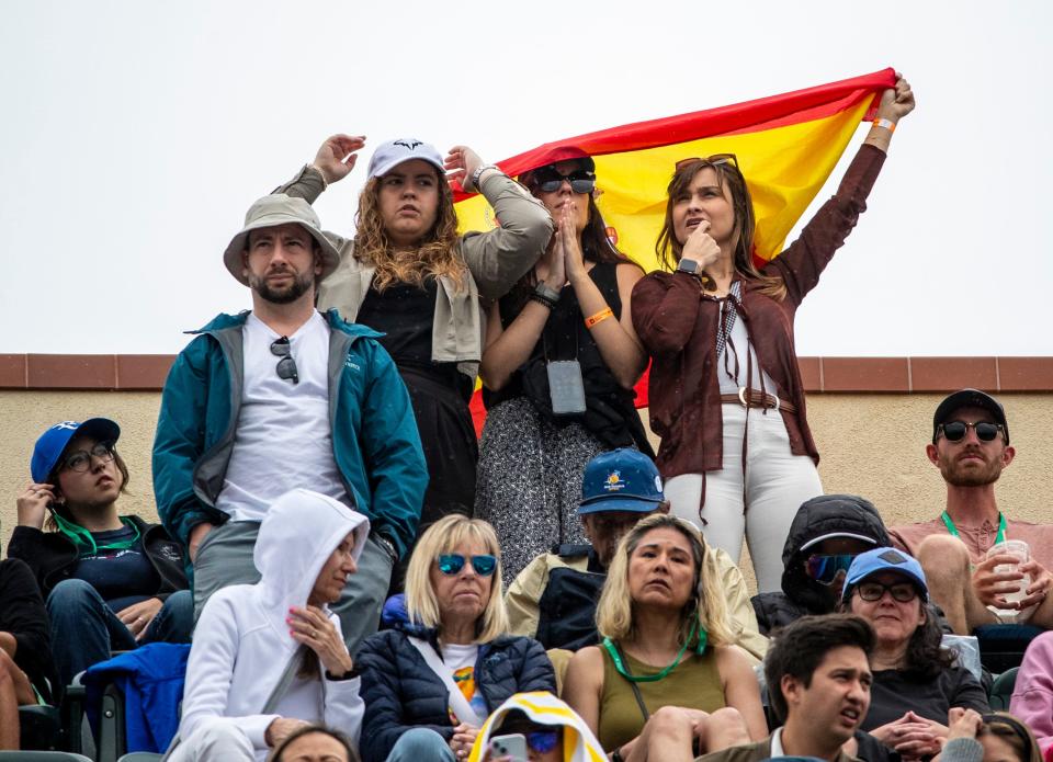A group of fans take shelter from rain under a Spanish flag during a rain delay in the ATP semifinal match between Carlos Alcaraz and Jannik Sinner at the BNP Paribas Open in Indian Wells, Calif., Saturday, March 16, 2024.
