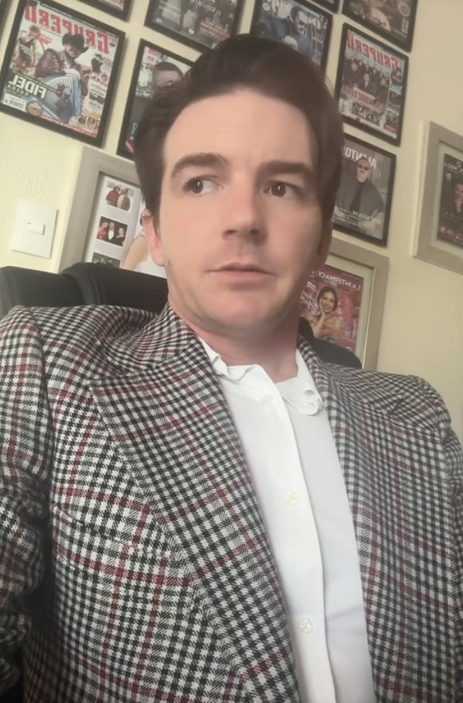 Drake in a houndstooth jacket, sitting, looking pensive with wall posters behind