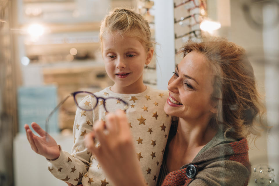 Happy mother helping her daughter with choosing new eyeglasses at optician store.