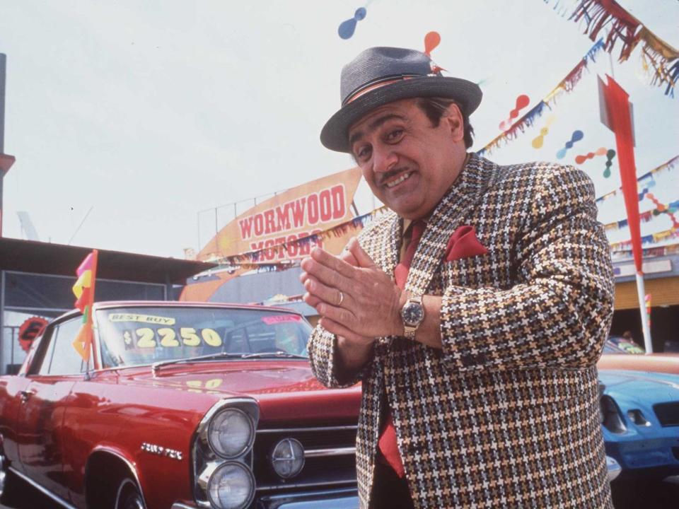 danny devito as harry wormwood in matilda. he's a greasy car salesman in a loudly pattnered jacket, red shirt, and red pocket square, wearing a grey hat. he's rubbing his hands together like he's about to con you