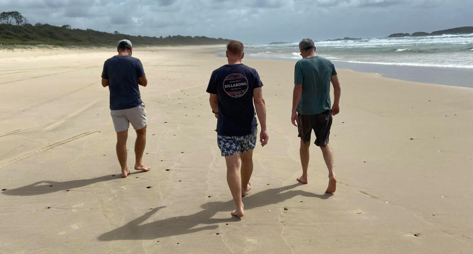 Three men, seen from behind, walk along a beach.