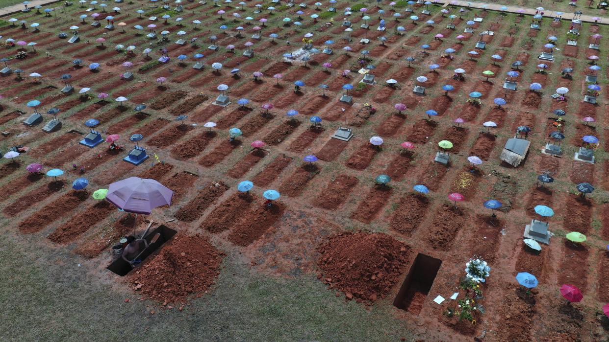 Shot from above, a worker digs a grave among hundreds of other recently dug gravesites, most marked with umbrellas of different colors.