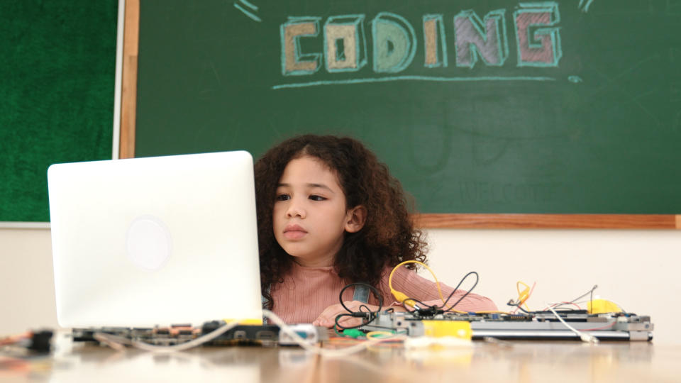 Young girl at a desk with a laptop and electronic components, with 'CODING' written on the chalkboard behind her