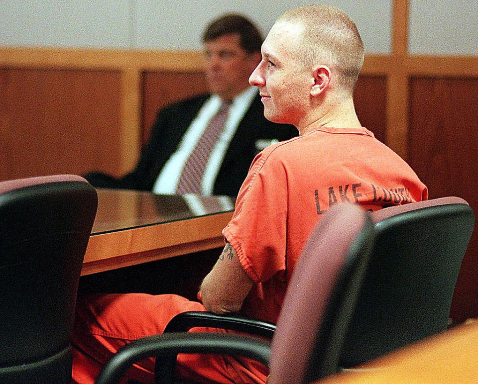 A man in a prison jumpsuit, sitting in a courtroom, looks forward. A suited man in the background is slightly blurred