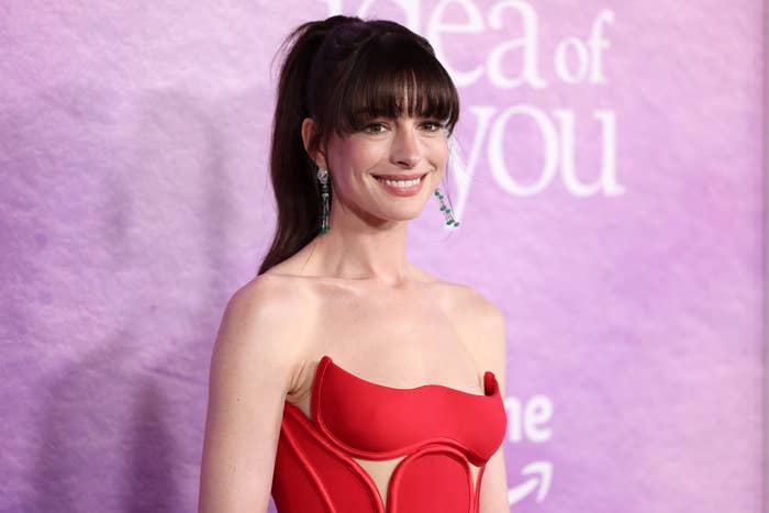 Anne Hathaway on the red carpet, wearing a strapless gown with a unique cutout design, smiling in front of a backdrop with text