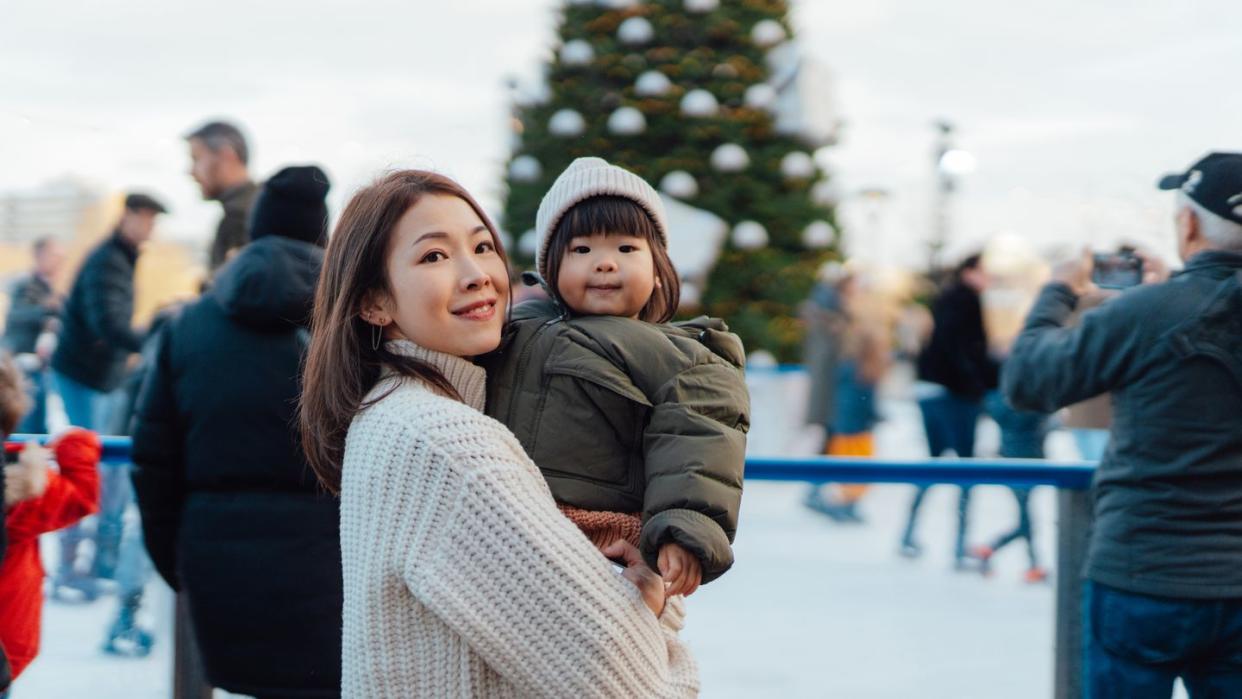 smiling young asian woman holding her cute baby girl, standing in front of the ice rink happy family having fun together at the amusement park christmas market