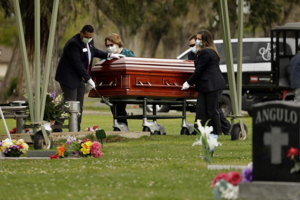 Four people in masks and gloves roll a casket at a cemetery.
