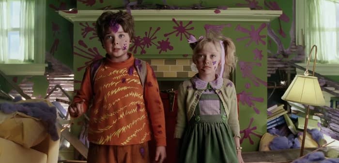 Two children are standing indoors with messy paint splatters on the walls and floors behind them. Both wear colorful, paint-stained clothes