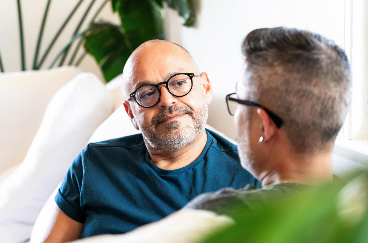 A man listens to his partner speak as they sit together face to face on their couch at home.