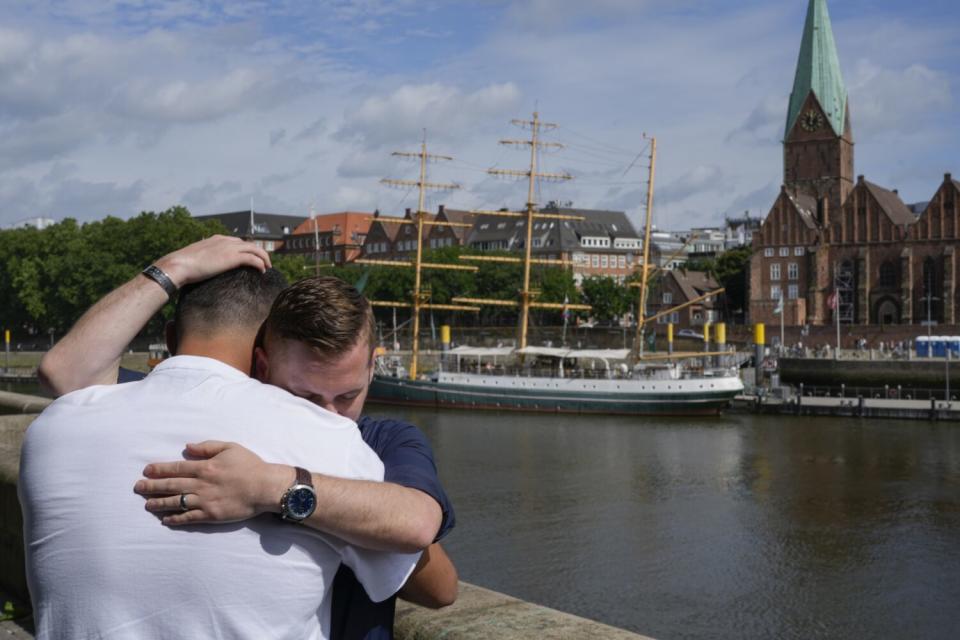 Two men embrace along a river with boats and buildings in the background