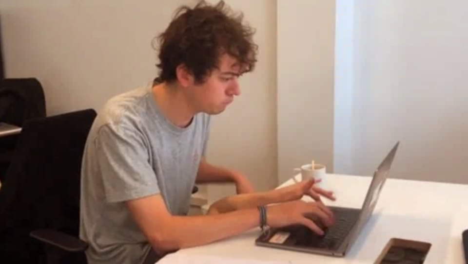 A person with curly hair sits at a table, looks at a laptop screen and types on the keyboard with one hand, while holding a disposable cup with the other.