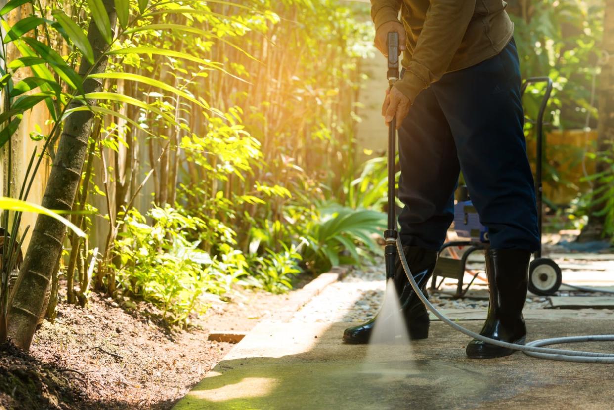 man in rain boots using hose to pressure wash concrete patio in yard with bright green sunlit bushes