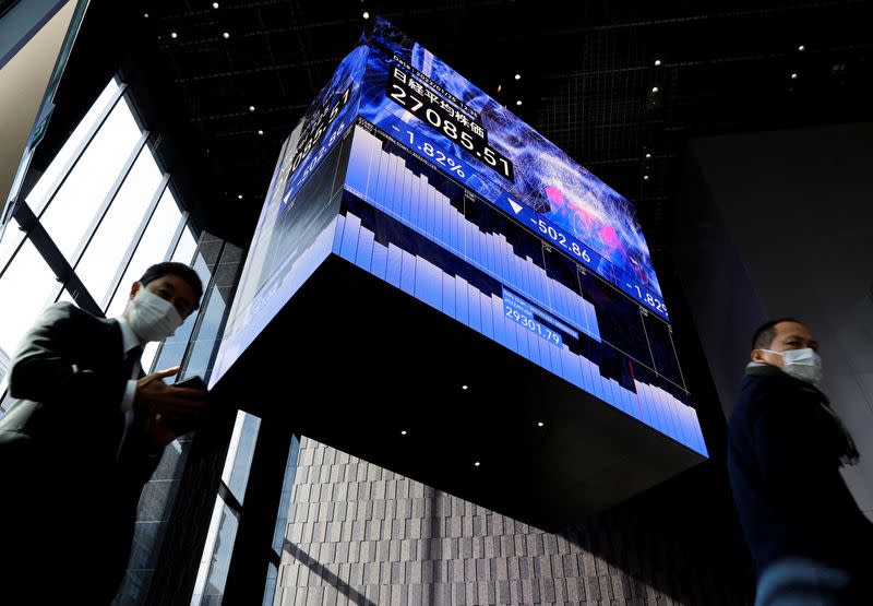 Men wearing protective face masks walk under an electronic board showing Japan’s Nikkei share average, amid the coronavirus disease (COVID-19) pandemic, in Tokyo