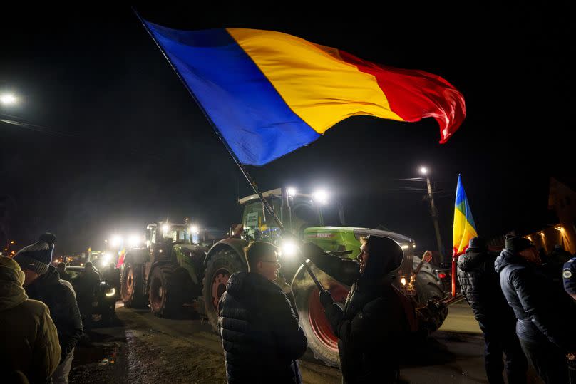 Ein Mann schwenkt eine Flagge neben einer Reihe von Traktoren, die die erste Spur einer Straße nach Bukarest blockieren, in Afumati, Montag, 15. Januar 2024.