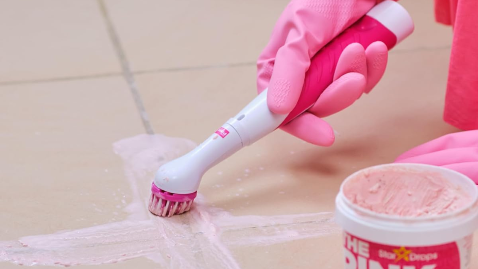 hand wearing a pink rubber glove using The Pink Stuff scrub brush to clean tiled floor grout