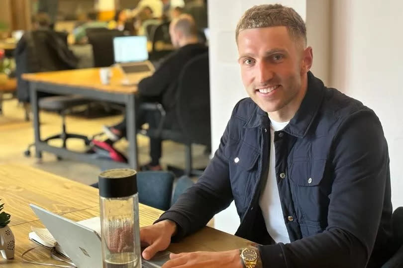 Man working on laptop in a co-working space- Tramshed Tech, Cardiff. He is sitting at the forefront of the image behind a desk with a water bottle, and other remote workers can be seen in the background using the space, on wooden tables in a large room which people can pay monthly memberships for or book out for the day. There are also facilities like a kitchen which can be spotted at the very back of the image.