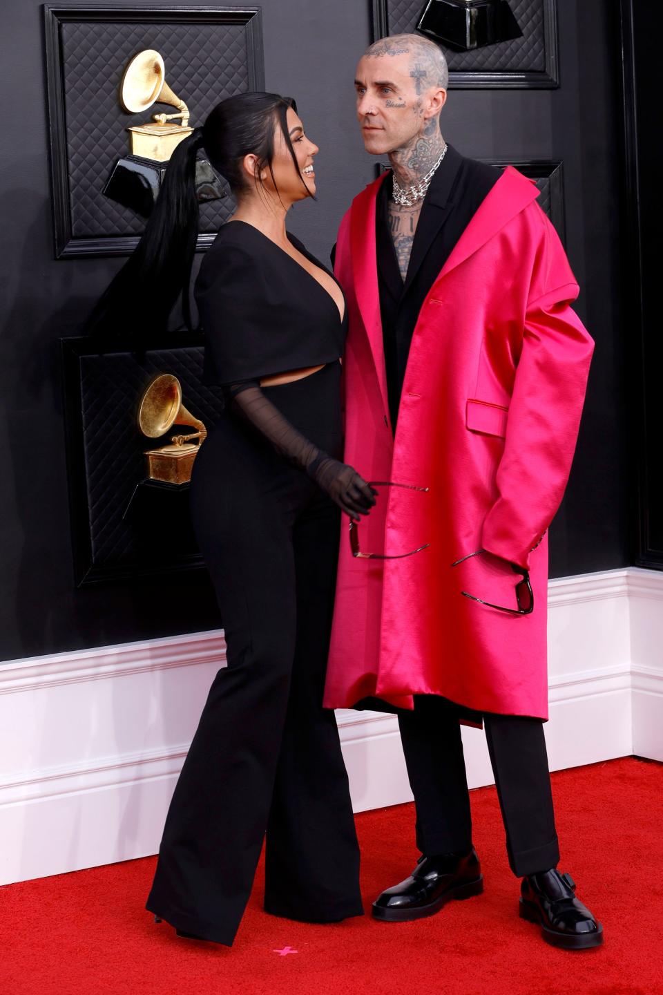 Kourtney in a black long-sleeve jumpsuit. Travis in a neon pink oversized coat over a black suit with no shirt and a crystal necklace.