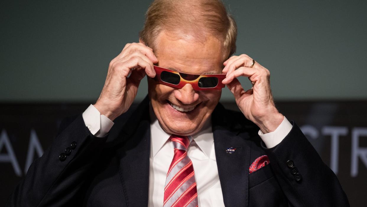  People wearing suits on a stage laugh as they hold paper eclipse glasses to their faces. 