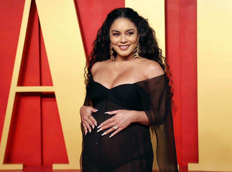 Vanessa Hudgens on the red carpet, wearing a sheer black dress that reveals her baby bump. She smiles with hands on her belly, accessorized with large earrings