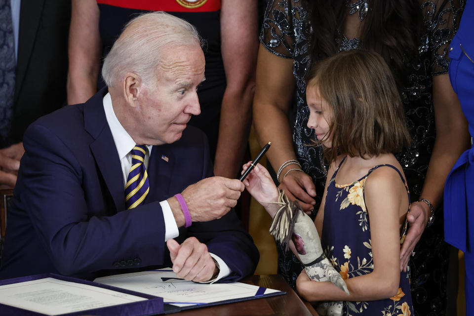 President Biden, seated at a desk, hands a pen to Brielle Robinson, standing and holding a plush toy.