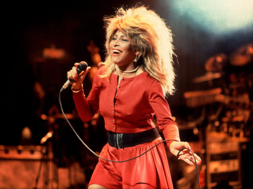 Tina Turner performing onstage while holding a microphone and wearing a red dress with a black strap around the waist.