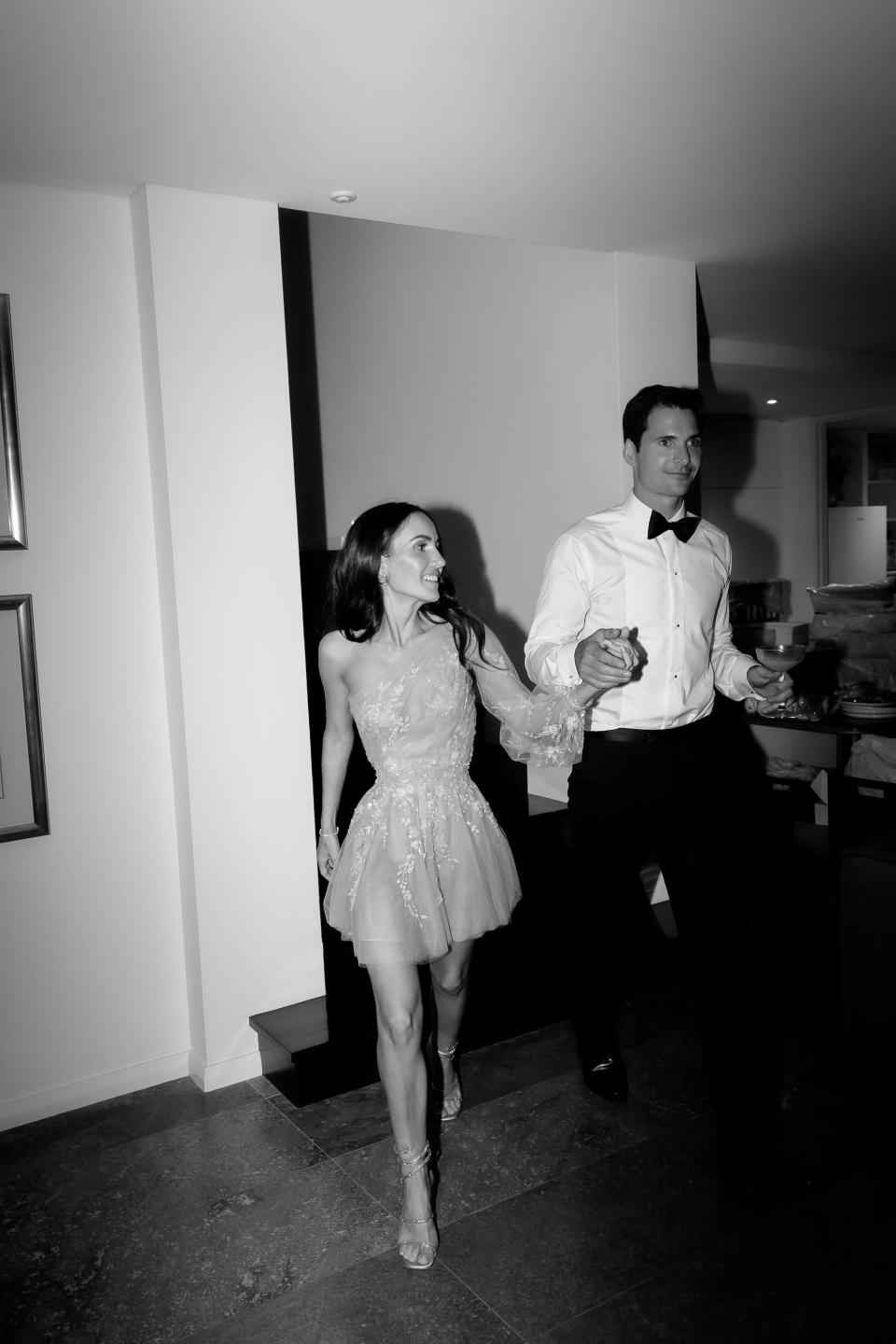 A bride and groom hold hands and walk through a room together.