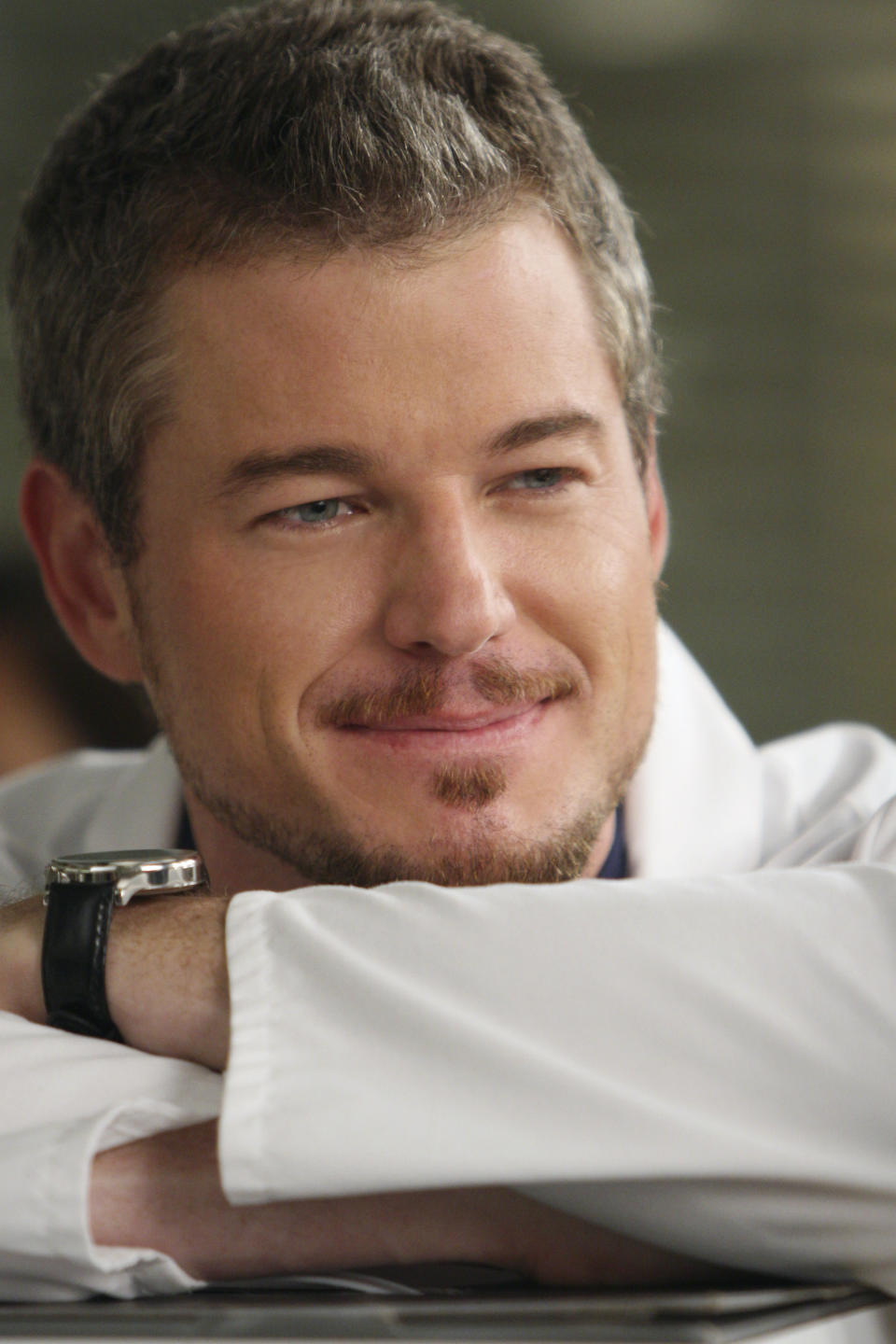 Eric Dane, in a reflective mood, rests his arms on a surface, smiling gently. He is wearing a white coat and a wristwatch