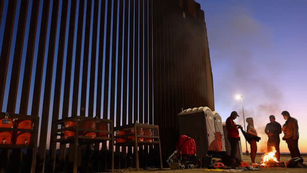 PHOTO: Immigrants from Cuba and Venezuela warm themselves by a fire before sunrise along the U.S.-Mexico border barrier as they await processing by the U.S. Border Patrol after crossing from Mexico on May 22, 2022 in Yuma, Ariz. (Mario Tama/Getty Images)