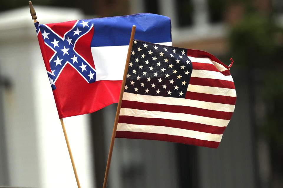 In this April 25, 2020 photograph, a small Mississippi state flag is held by a participant during a drive-by "re-open Mississippi" protest past the Governor's Mansion, in the background, in Jackson, Miss. This current flag has in the canton portion of the banner the design of the Civil War-era Confederate battle flag, that has been the center of a long-simmering debate about its removal or replacement. (AP Photo/Rogelio V. Solis)
