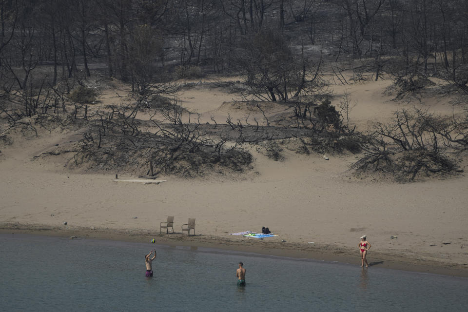 FILE - People play with a ball in front of a burnt forest at a beach of Glystra, on the Aegean Sea island of Rhodes, southeastern Greece, on July 27, 2023. Tourists at a seaside hotel on the Greek island of Rhodes snatched up pails of pool water and damp towels as flames approached, rushing to help staffers and locals extinguish one of the wildfires threatening Mediterranean locales during recent heat waves.(AP Photo/Petros Giannakouris, File)