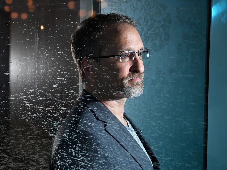 A man with grey hair, beard, and glasses stands behind a glass wall staring off into the distance.