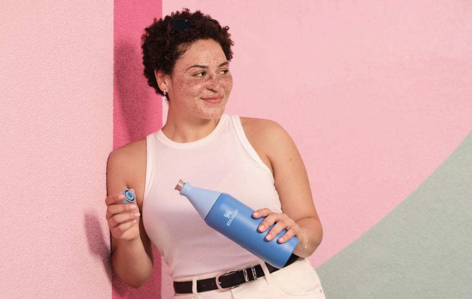 A woman stands against a pink wall holding a blue Stanley All Day Slim Water Bottle. She has a smile on her face as she looks to the side. 