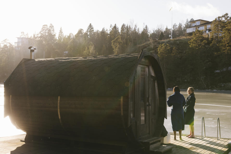 Two people in robes stand by a barrel sauna near a body of water with trees and a house in the background