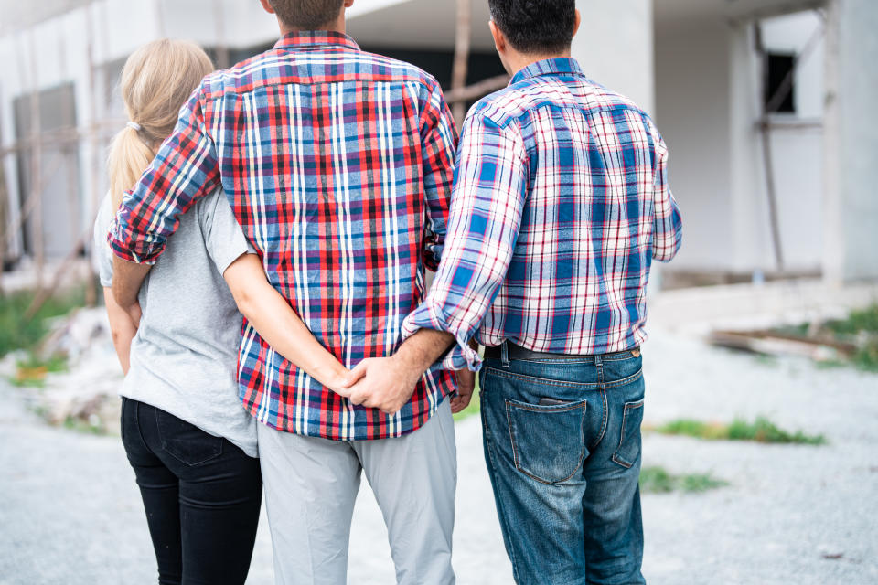 Three people from behind, one woman and two men in casual clothes, arms around each other, with the woman subtly holding hands with one man behind the other’s back