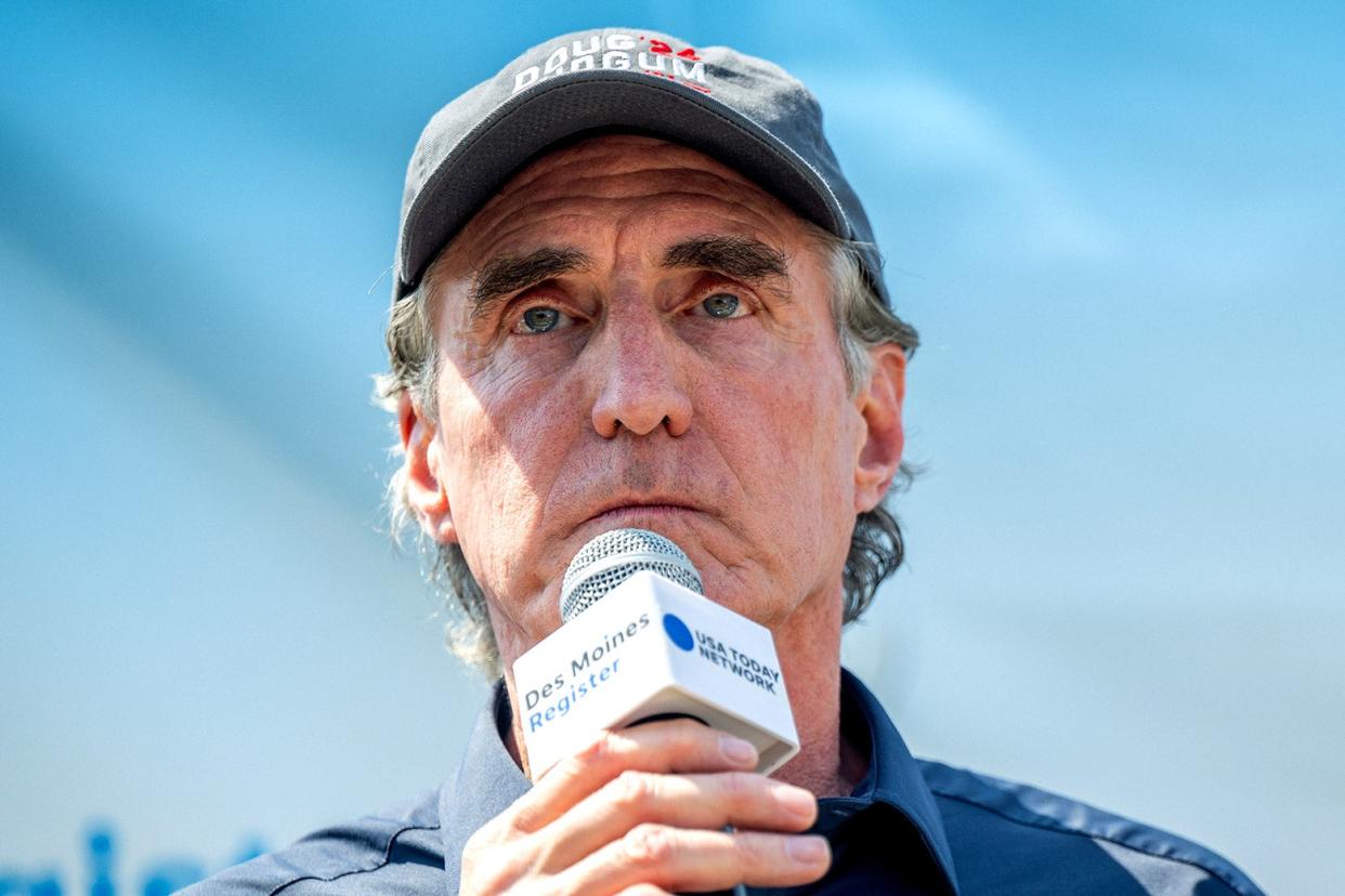 A close-up photograph of a man with a microphone wearing a baseball cap.