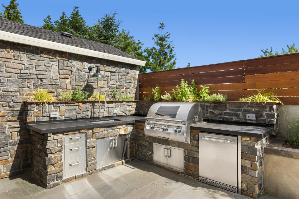 Outdoor kitchen with stone countertops, built-in stainless steel grill, sink, and storage, surrounded by stone and wood walls with plants growing on top