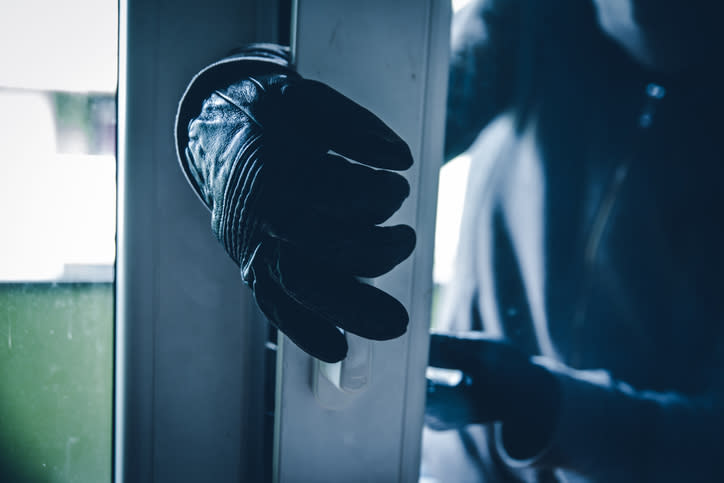 A gloved hand opening a glass door, depicting a potential burglary or break-in scenario. The scene suggests a sense of urgency and caution