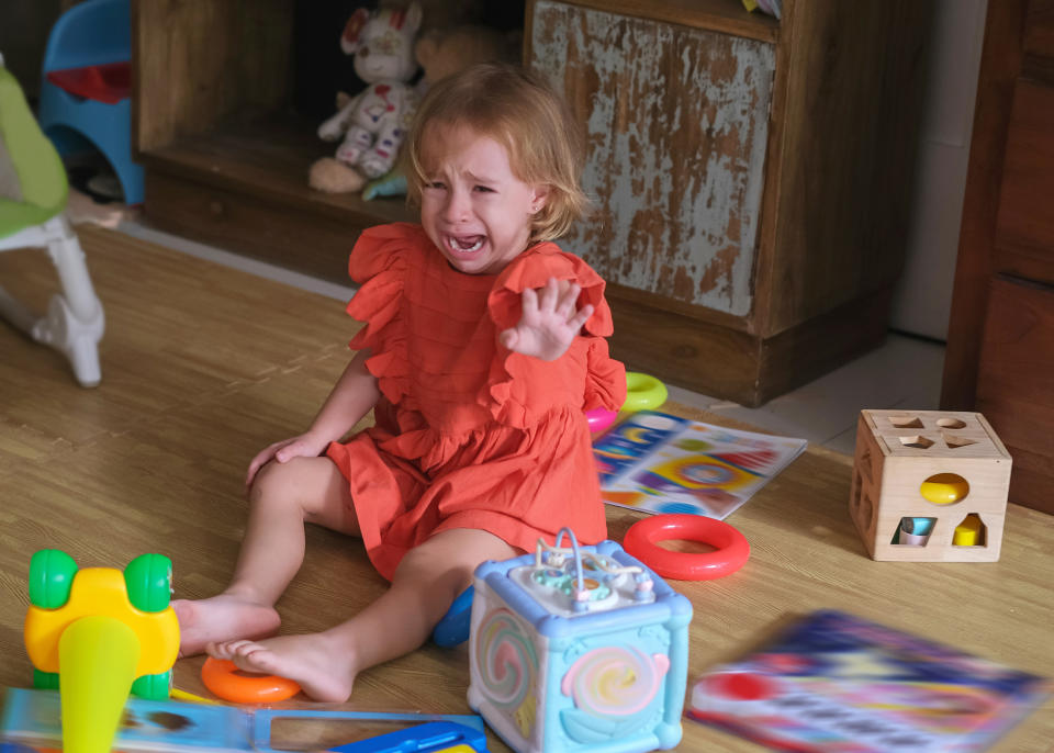 A young child in a ruffled dress cries on the floor amidst scattered toys