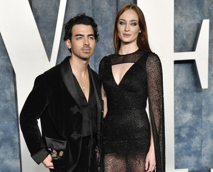 Joe Jonas in a black velvet suit posing next to Sophia Turner in a sheer v-neck black gown