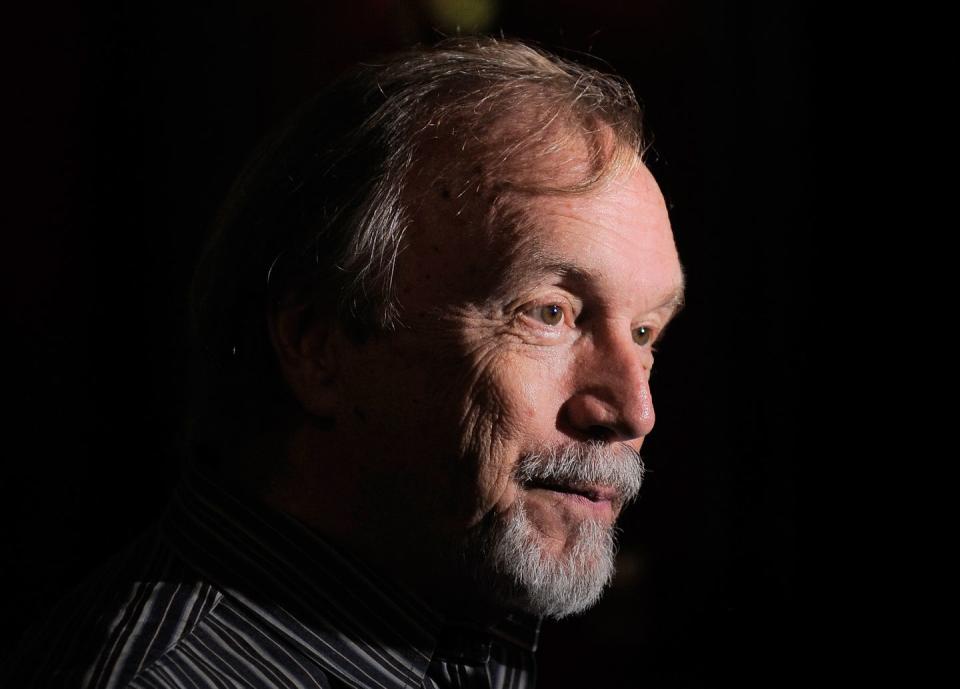 a closeup of george lee andrews in a dark room, wearing a striped black shirt, looking off camera