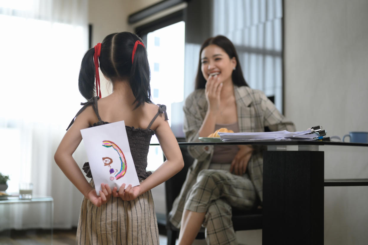 Cute little girl hiding greeting card for mother behind back.