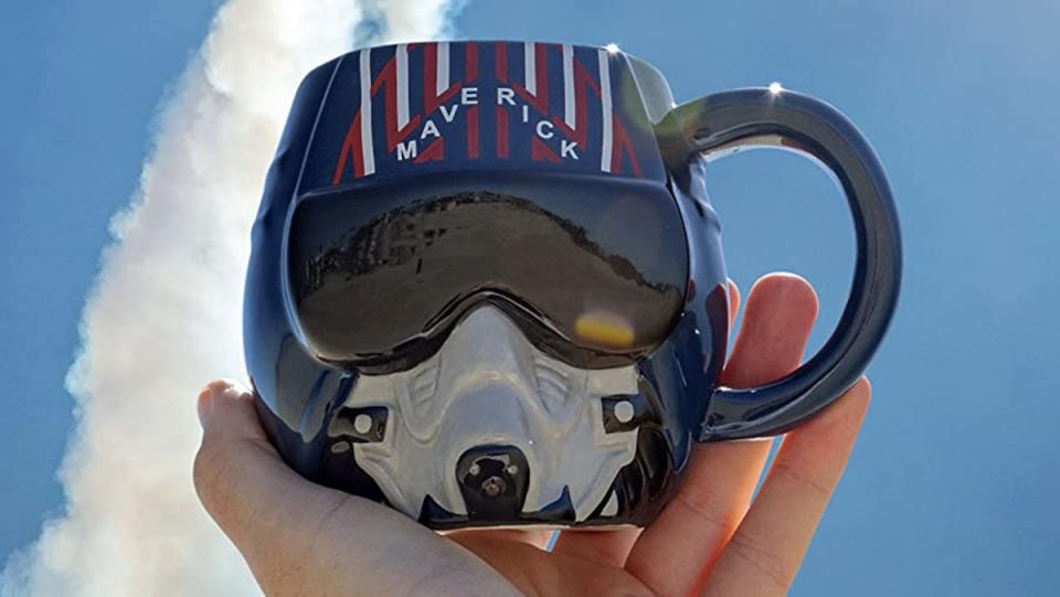 A hand holds up a ceramic mug of a navy pilot helmet from Top Gun: Maverick against a blue sky with jet flumes