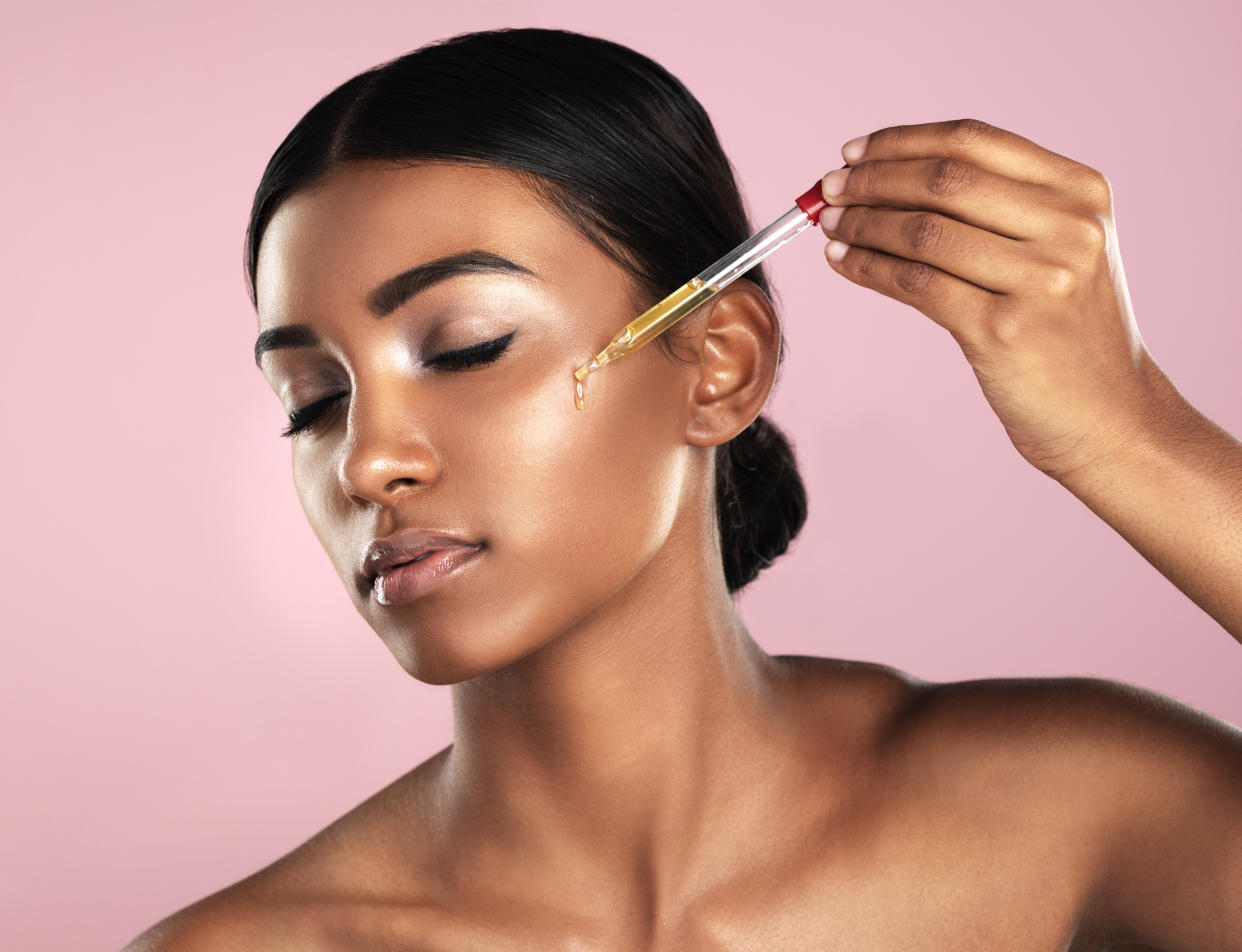 Studio shot of a beautiful young woman applying essential oil to her face with a dropper posing against a pink background