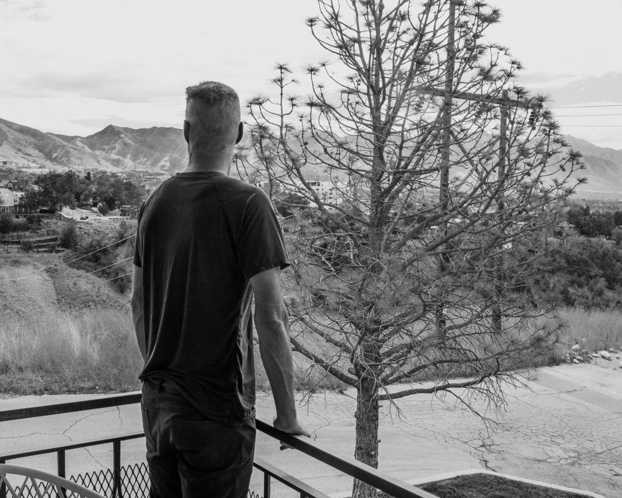 A black and white image of a man in a black t-shirt facing away from the camera, leaning on a balcony railing looking out towards trees and mountains.