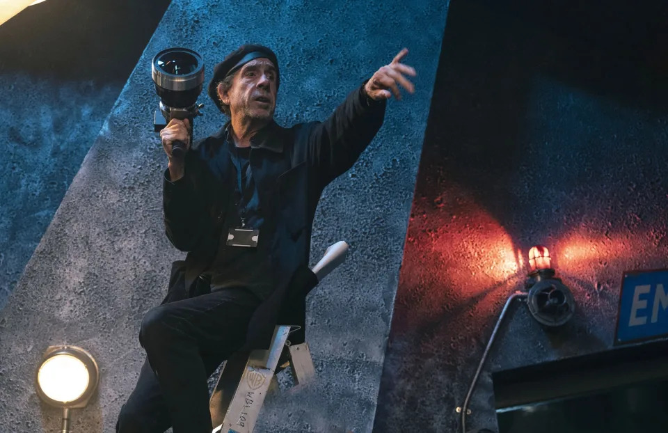 A film director wearing a beret and holding a camera lens points while perched on a ladder