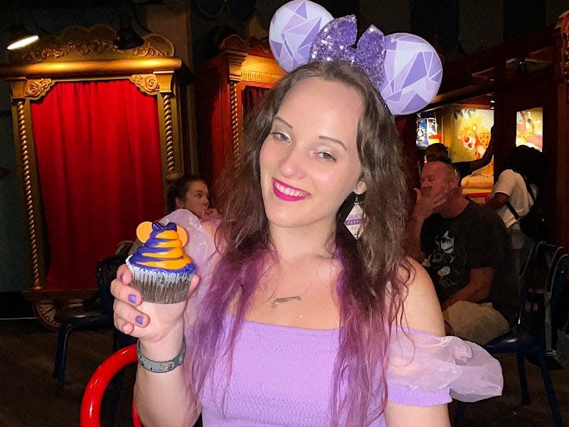 The writer wears purple mouse ears and a purple shirt and holds a purple-and-orange cupcake for Halloween