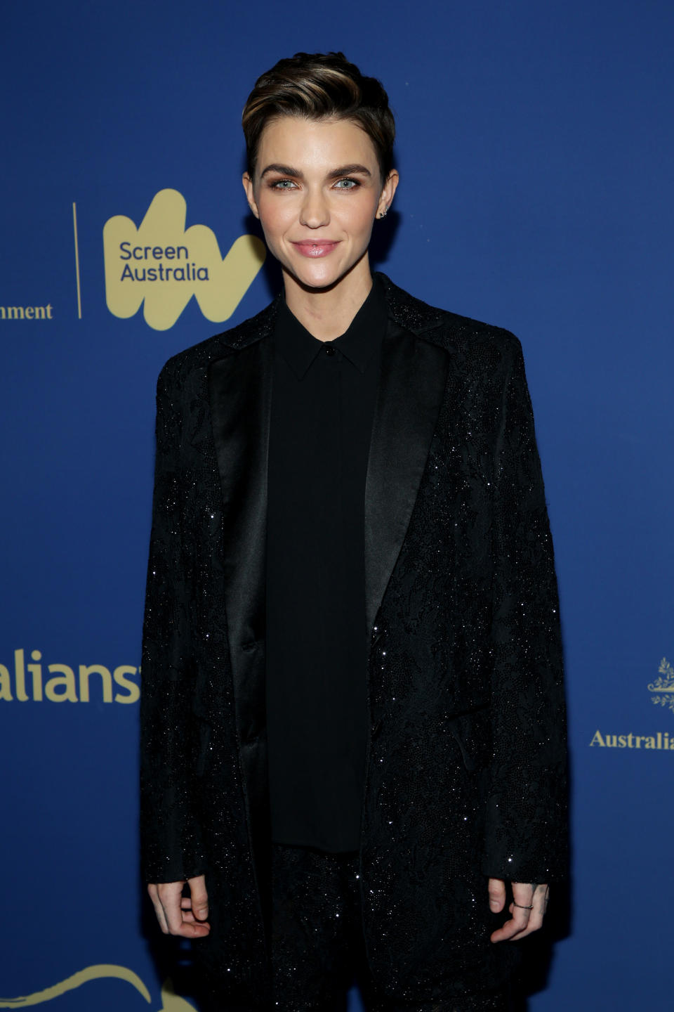 Ruby Rose in a black glittery suit and matching shirt, posing at an event with a Screen Australia background