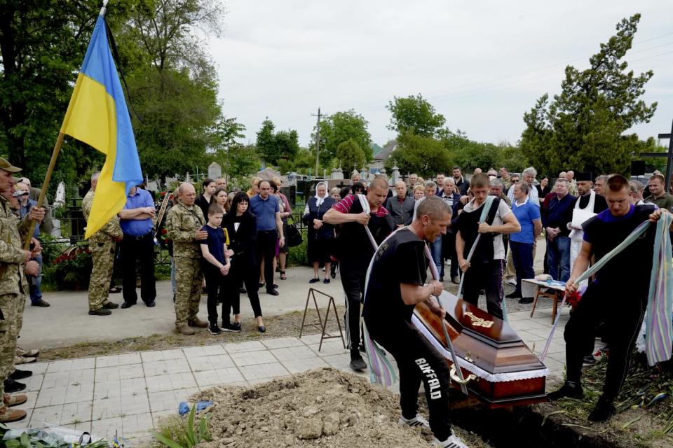 Four people in dark clothing use long pieces of fabric as ropes to lower a coffin into a grave as other people watch