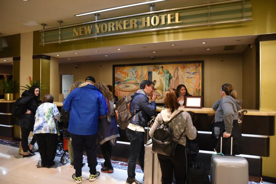 A shot of the check-in desk at the New Yorker Hotel, which has an Art Decor painting behind the workers standing at the desk, and gold letters that spell out New Yorker Hotel above. Tourists in jackets with backpacks and suitcases stand in front of the desk.