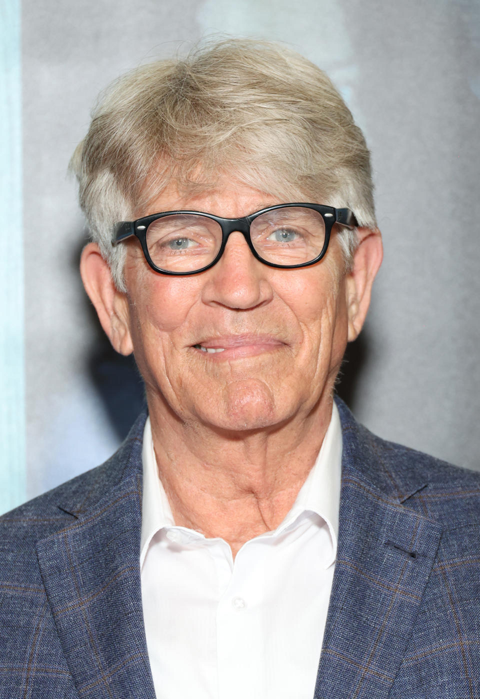 Eric Roberts smiling, wearing black-framed glasses, a white shirt, and a checkered blazer